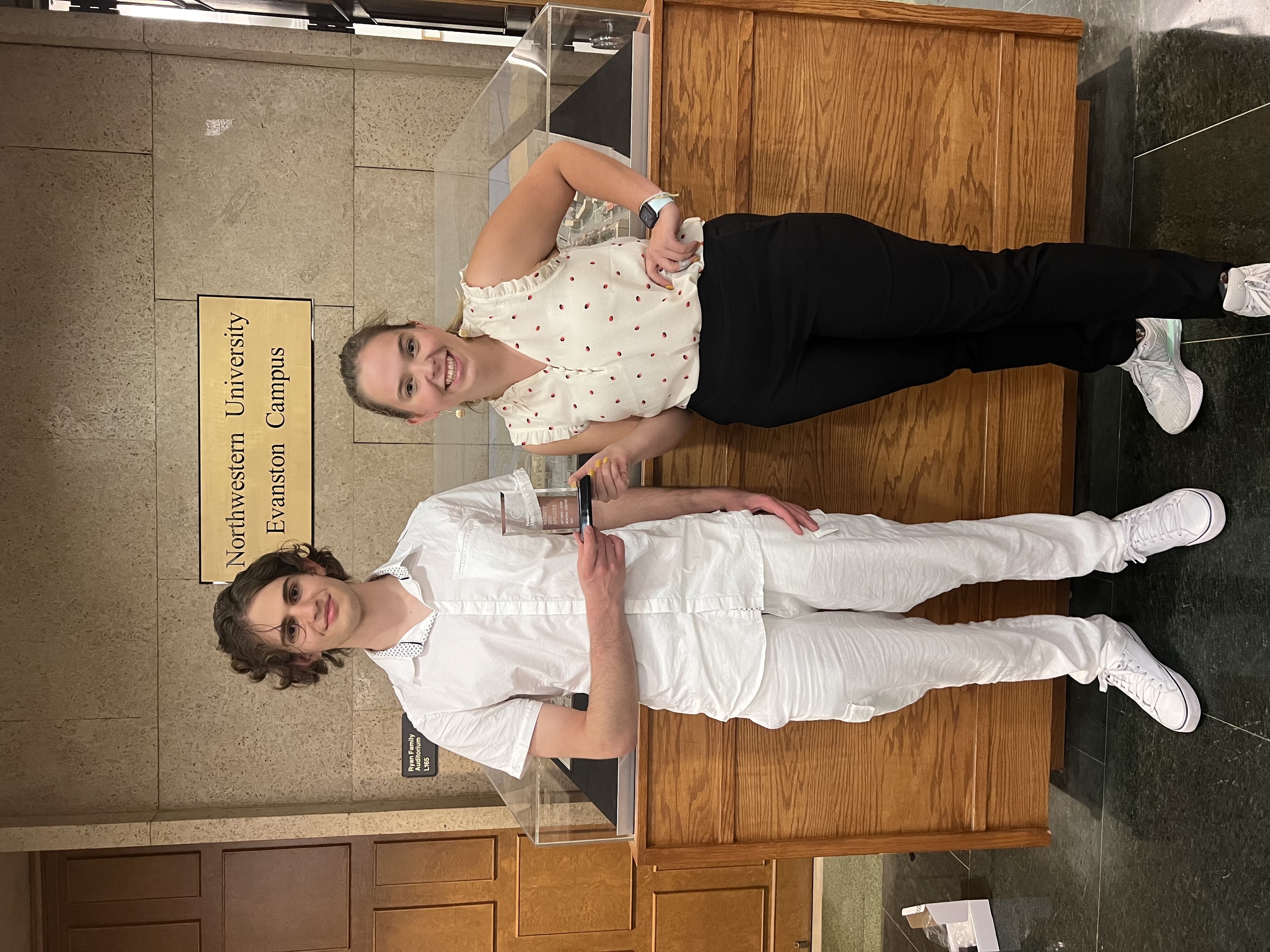 Two debaters one wearing all white, the other wearing a white blouse and black pants, both holding a glass trophy. 