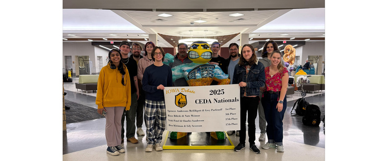 A group of smiling debaters and coaches standing around a stature of herky holding a banner 