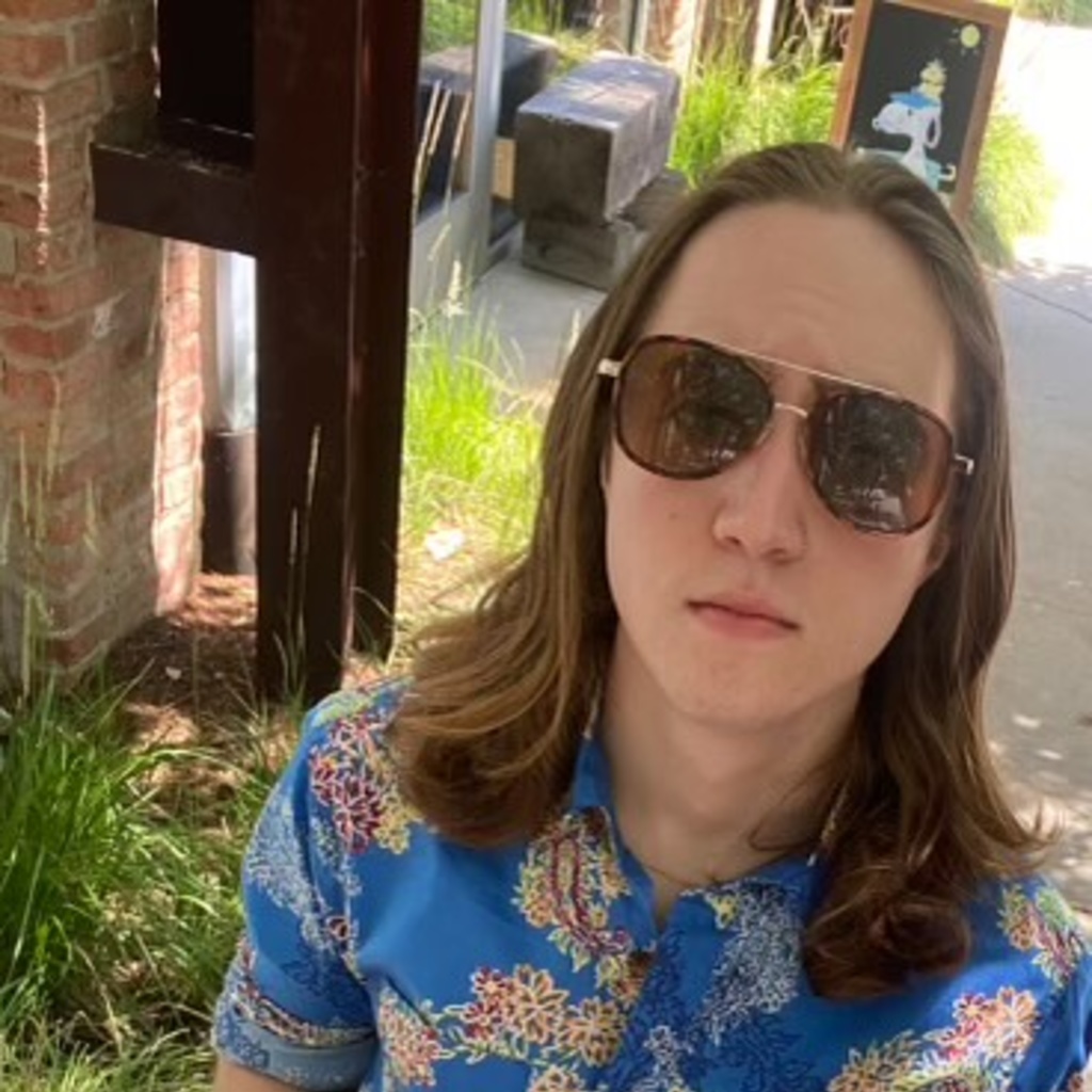 A white person with long brown hair wearing sunglasses and a blue floral suit 