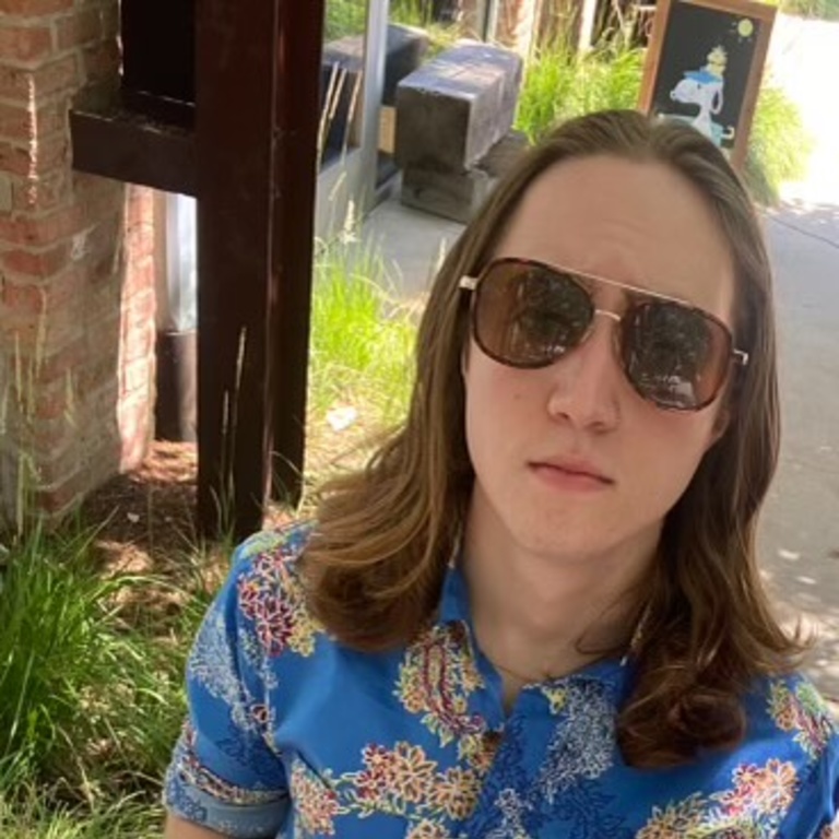 A white person with long brown hair wearing sunglasses and a blue floral suit 