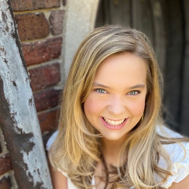 A smiling white women leaning agasint a wall. She has blonde hair and a white shirt 