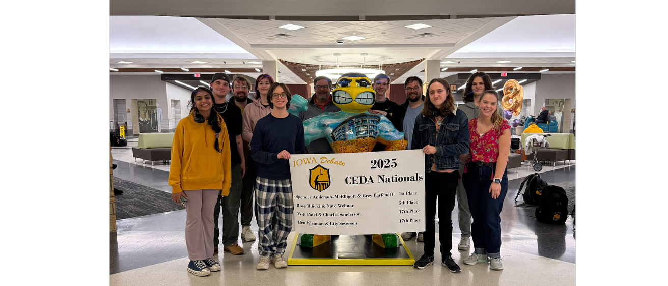 A group of smiling debaters and coaches standing around a stature of herky holding a banner 